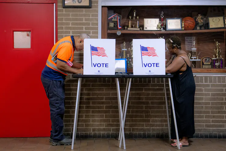 A man in an orange safety shirt and a woman in a long black dress each stand behind signs that say 'vote.'
