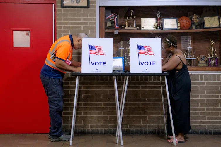 A man in an orange safety shirt and a woman in a long black dress each stand behind signs that say 'vote.'