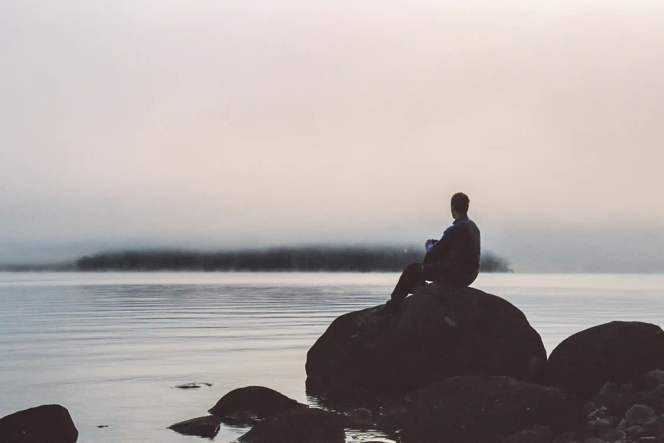 A man seen in silhouette sits atop a large rock looking out over the ocean on a gray day.