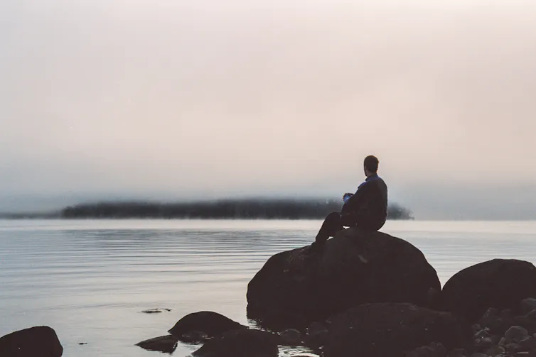 A man seen in silhouette sits atop a large rock looking out over the ocean on a gray day.