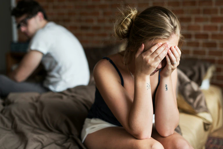 A caucasian heterosexual couple sitting at either ends of a bed while the woman rubs her forehead and the man looks back at her.