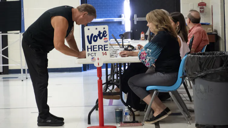 A man leaning over a table where quite a few people sit, with a sign 'Vote HERE' next to the table.