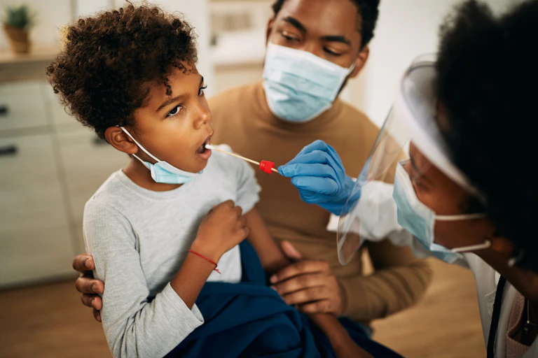 A child receives a swab to test for covid. He is being held by his father, who is wearing a face mask. A female health worker is performing the swab. She is also wearing a face mask.