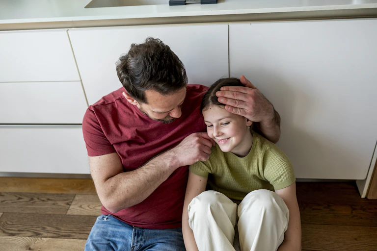 A man in a red T-shirt sits on the floor beside a young girl, with his arm around her and one hand resting gently on her head. The girl smiles as they sit close together.