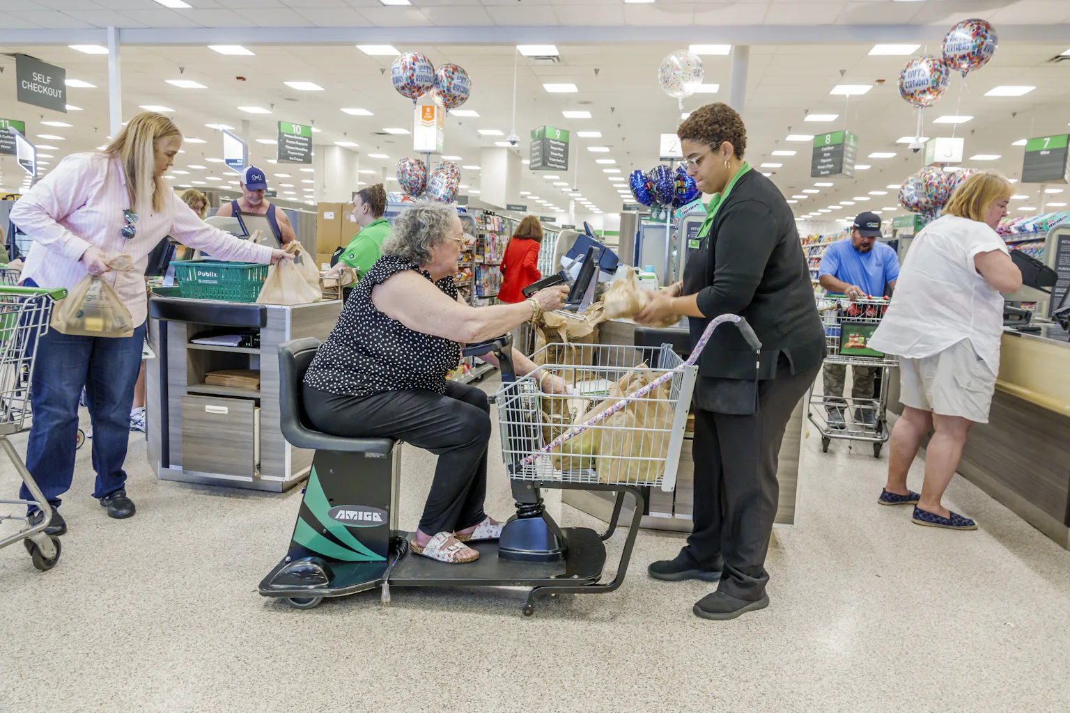 A woman idling an electric mobility grocery cart pays a cashier for groceries in a supermarket.