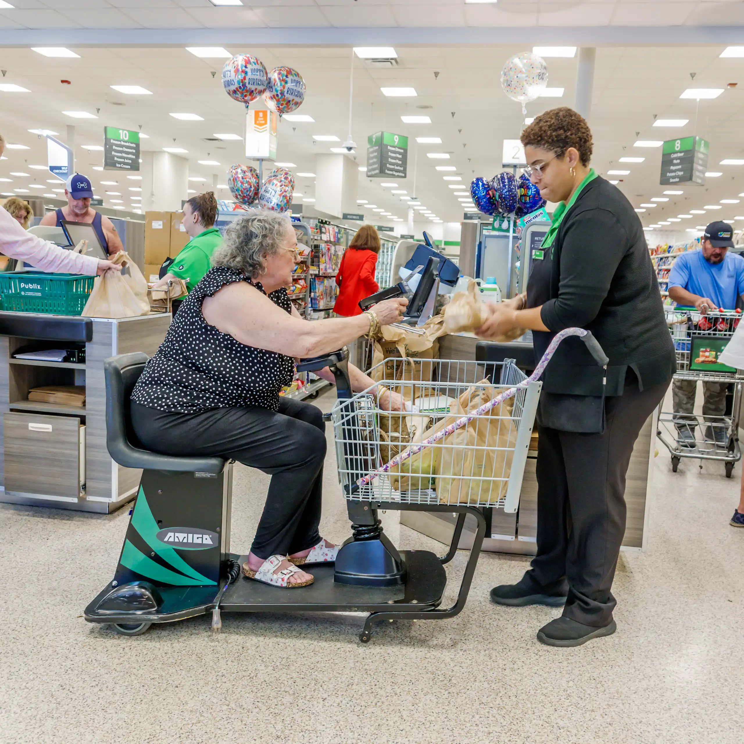 A woman idling an electric mobility grocery cart pays a cashier for groceries in a supermarket.
