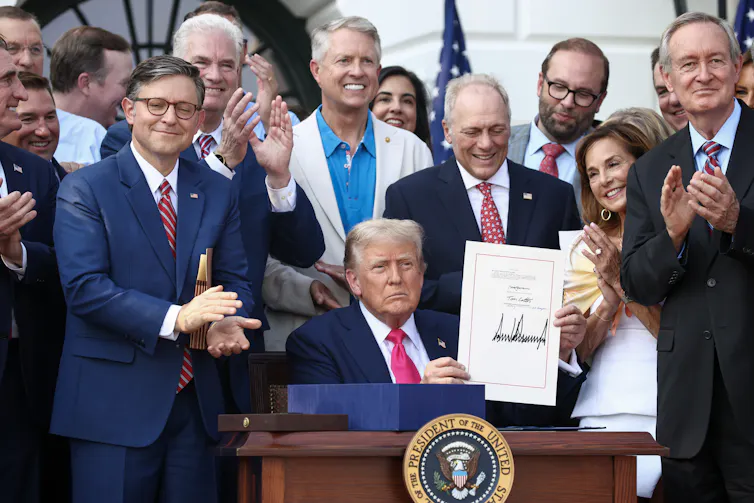 A man sitting at a desk holds up a piece of paper while surrounded by other people who are smiling.