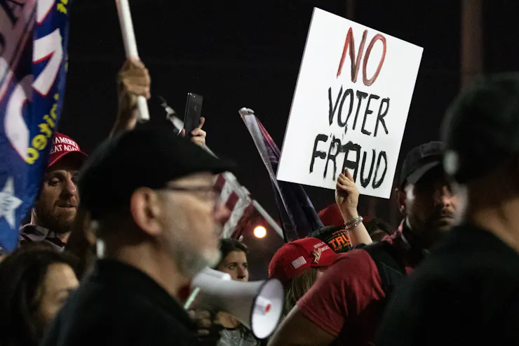A person in a crowd indoors holds a white sign with the words 'No Voter Fraud' on it.