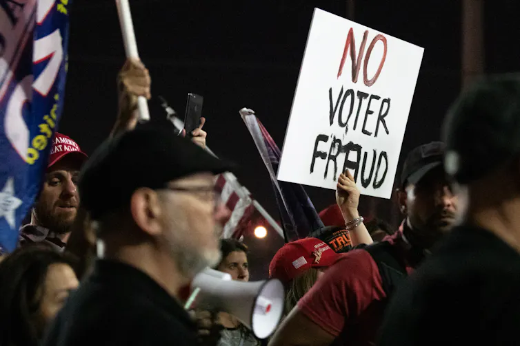 A person in a crowd indoors holds a white sign with the words 'No Voter Fraud' on it.