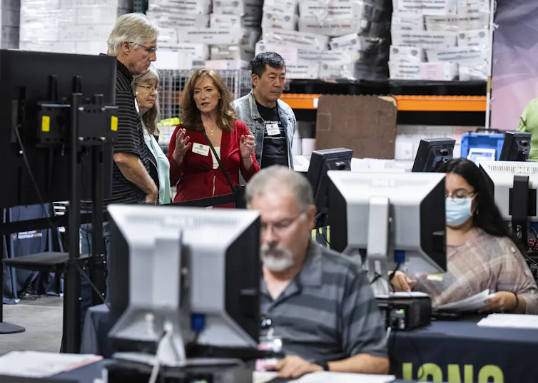 A woman talks to a man in an office while other people work in front of computers.
