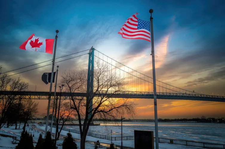 A silhouette of a bridge with U.S. and Canadian flags flying on flagpoles.