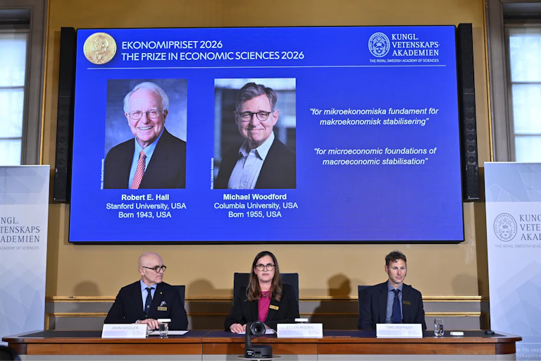 nobel panel sitting in front of a board showing the pictures of economics laureates robert e hall and michael woodford.
