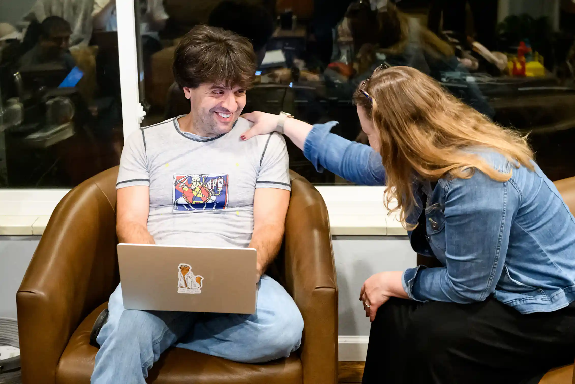 Karl Deisseroth sitting and smiling in a chair with laptop in his lap, Michelle Monje-Deisseroth sitting adjacent to him reaching a hand out to hold his shoulde