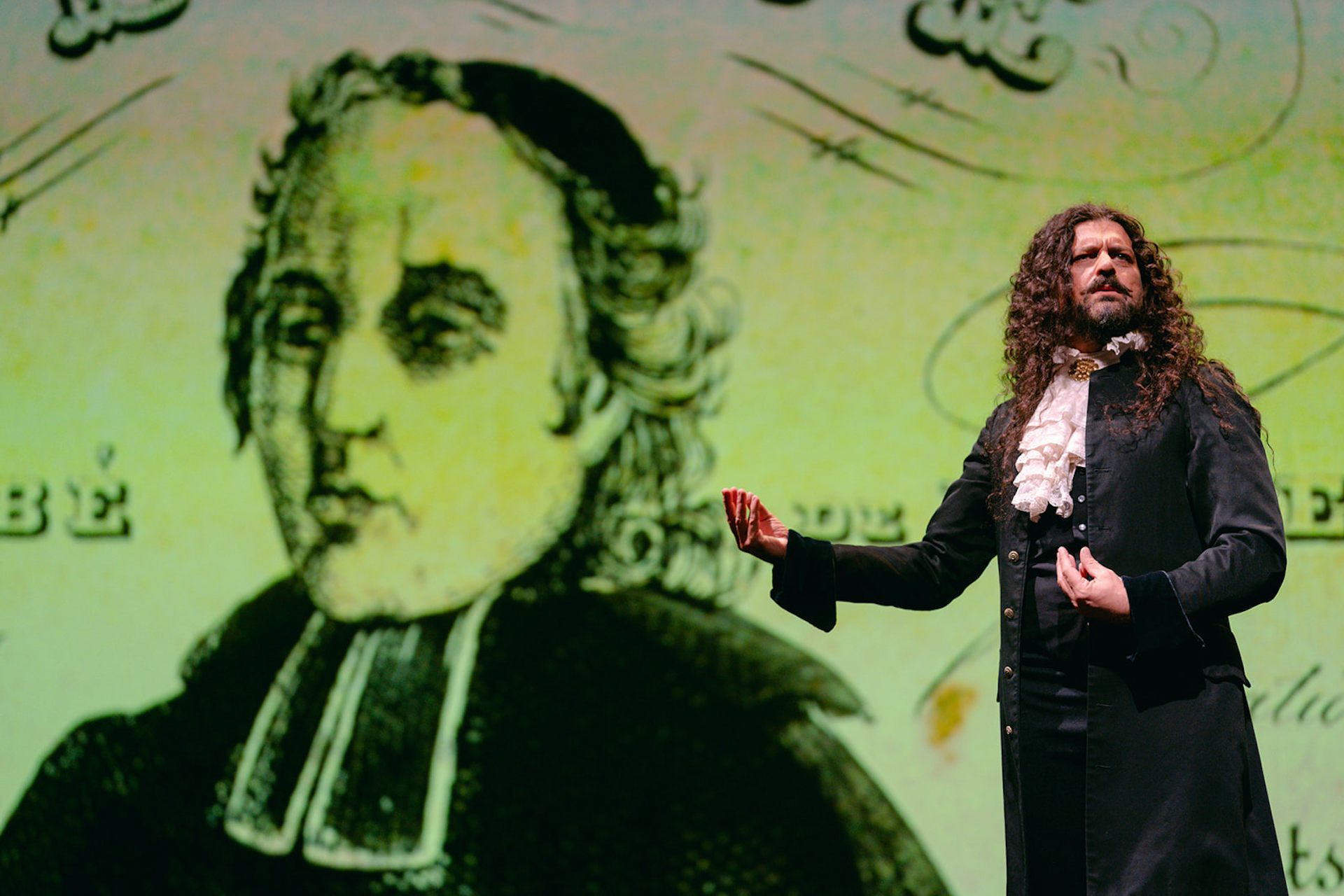 A man with long curly hair wearing a 19th century suit and frilled cravat stands in front of a screen with an illustration of a clergyman on it.