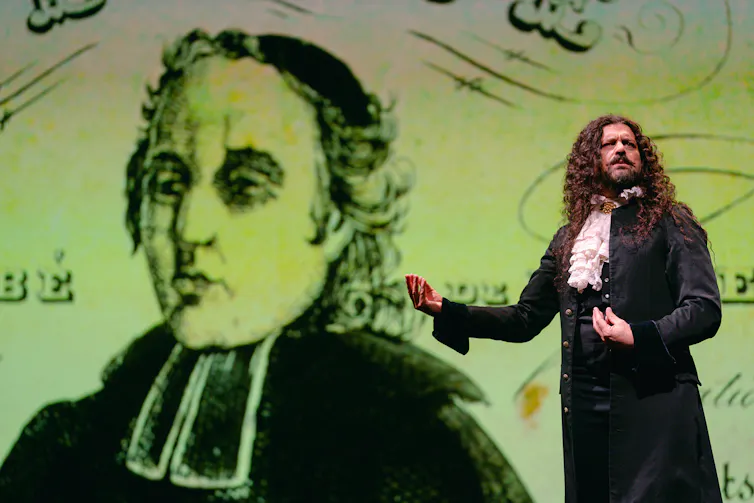 A man with long curly hair wearing a 19th century suit and frilled cravat stands in front of a screen with an illustration of a clergyman on it.