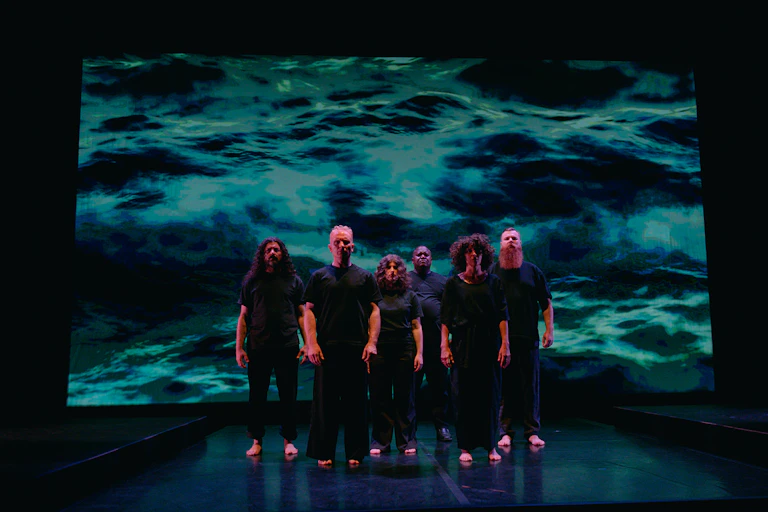 Six performers stand in a close cluster on a darkened stage, with an image of water projected on a screen behind them.