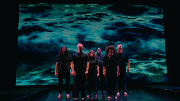Six performers stand in a close cluster on a darkened stage, with an image of water projected on a screen behind them.
