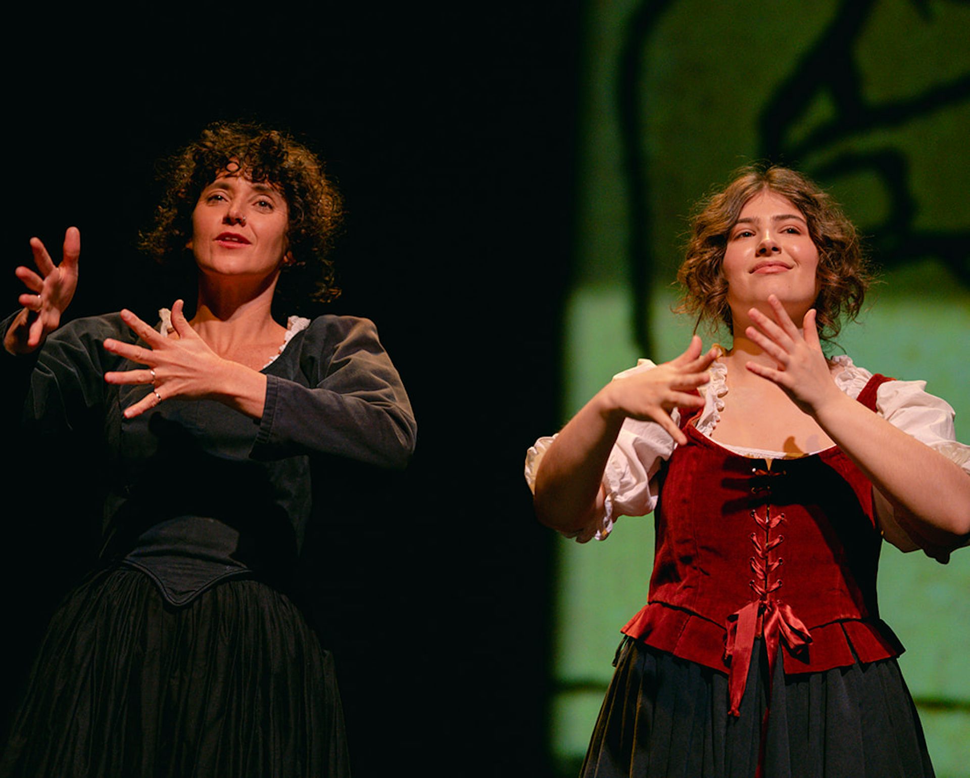 wo women in historical costumes stand on stage gesturing with their hands.