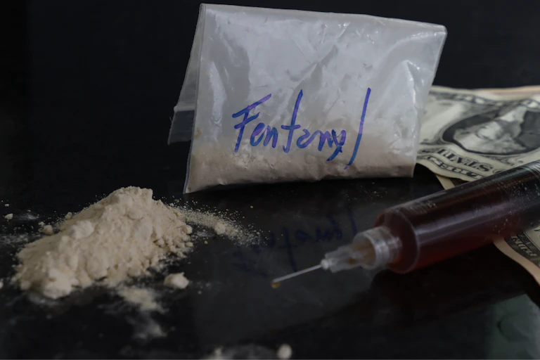 Dollar bills, a fentanyl powder pack and a syringe on a dark table.