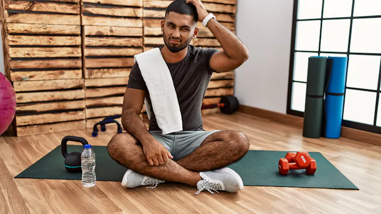 A confused man scratches his head while sitting on an exercise mat. He has a bottle of water, a kettlebell and two dumbbells sitting beside him.