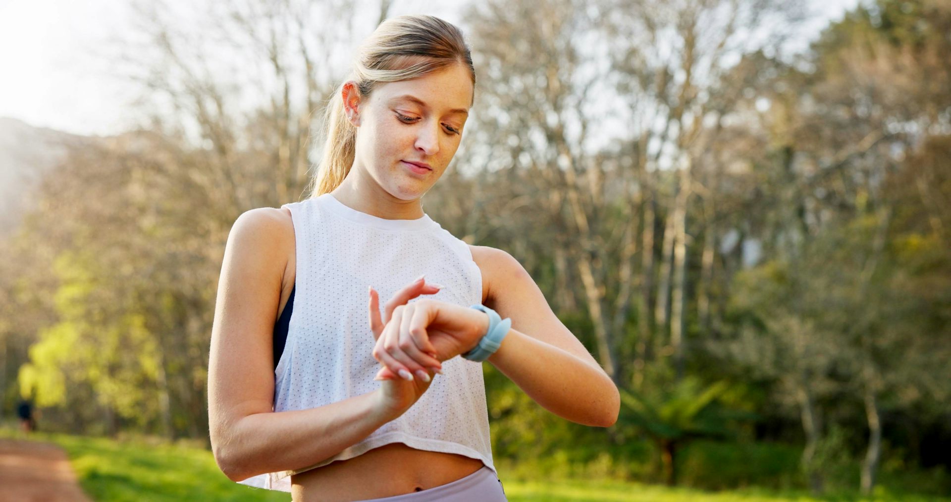 A woman standing outdoors in workout gear looks at her smartwatch.