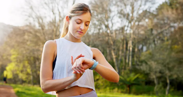 A woman standing outdoors in workout gear looks at her smartwatch.