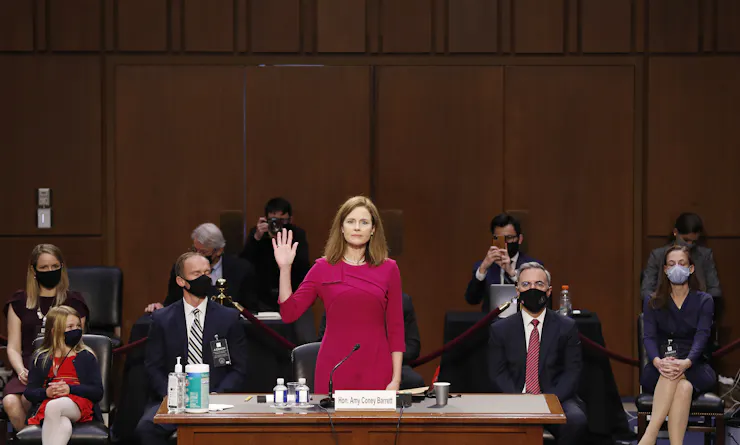 A woman in a burgundy dress stands in front of a table and raises her right hand while several people wearing masks look on.