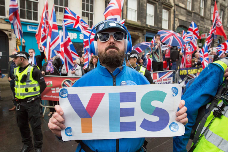 scottish independence supporter holding a yes banner while a crowd waving union flags stand behind him.
