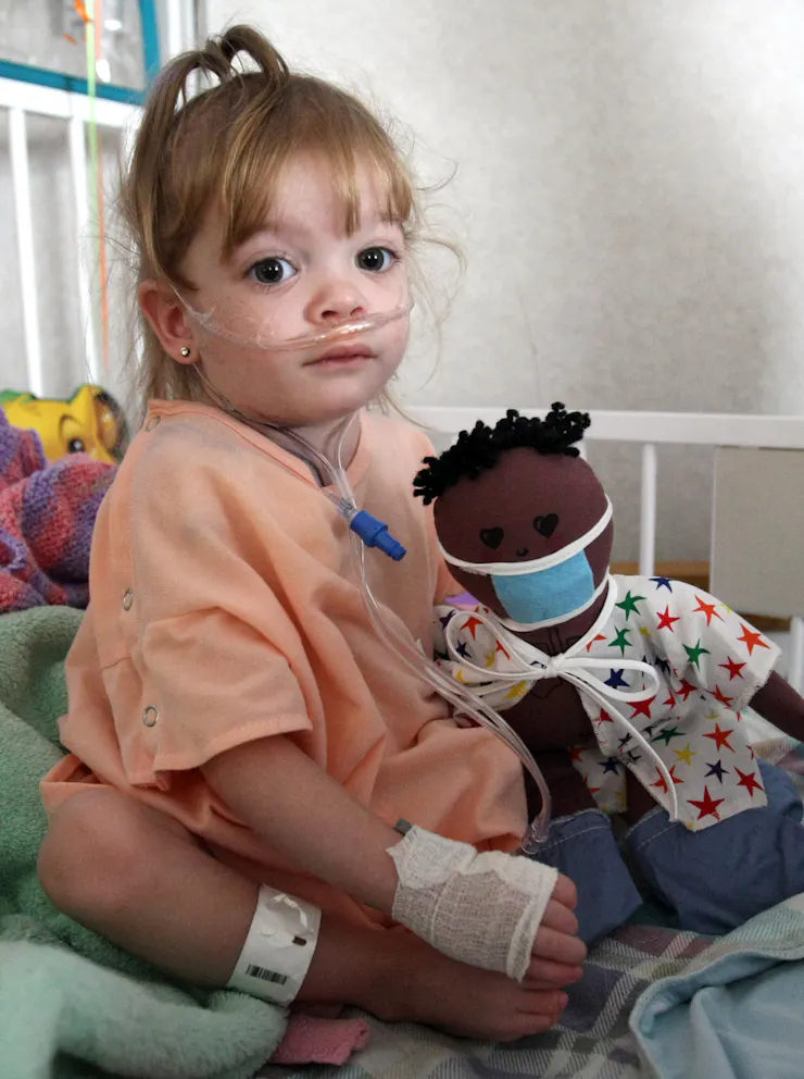 Child with nasal cannula sitting on bed, holding a doll wearing a surgical mask