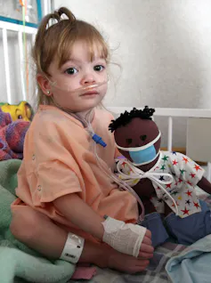 Child with nasal cannula sitting on bed, holding a doll wearing a surgical mask