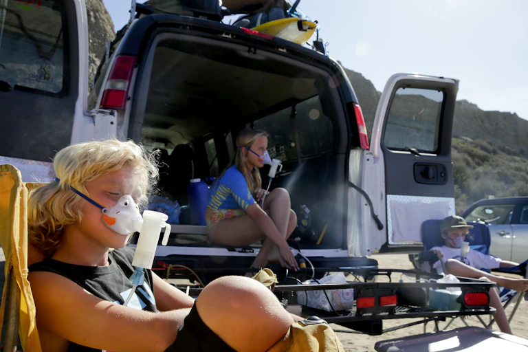 Three children sitting in beach chairs and the back of a van at the beach, wearing nebulizers on their faces