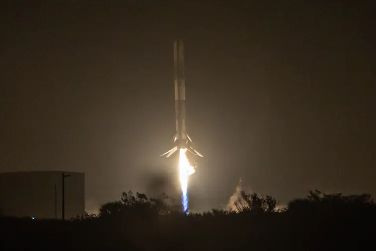 A photo of a rocket booster, that looks like a long cylinder with a small flame coming out of its end, landing in the dark.