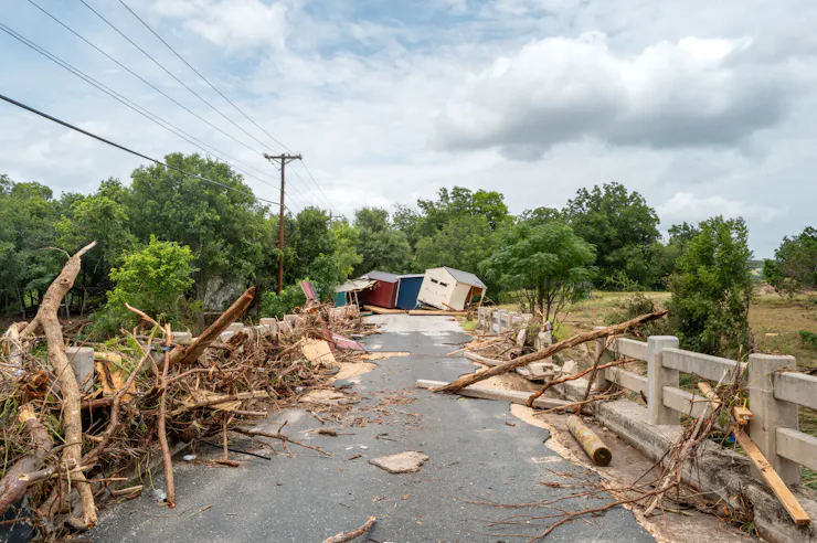 Flood debris has been pushed to the side of a road with two buildings off kilter at the end.