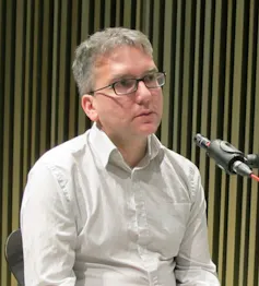 A photograph of Mark Fisher's face and torso, clad in a white shirt. He is at a microphone.