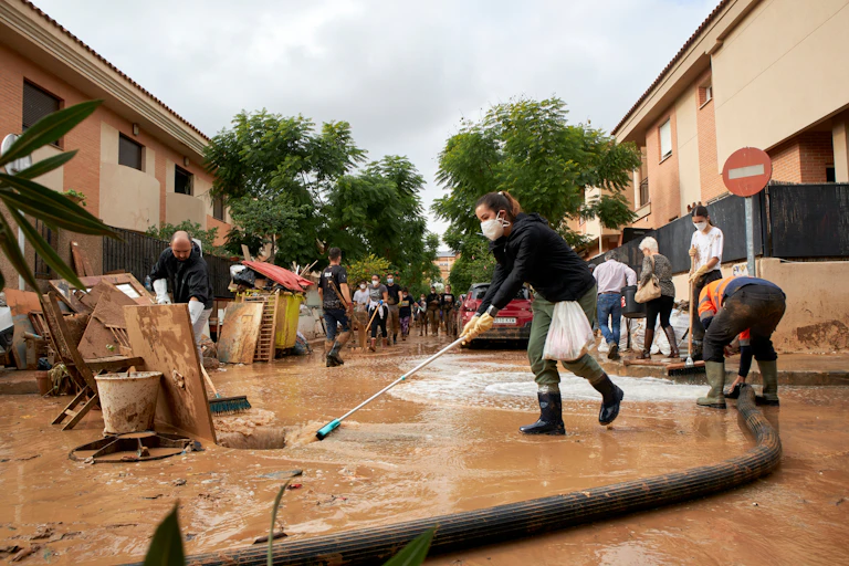 Un grup de persones amb mascareta, guants i botes neteja l'aigua i el fang d'un carrer.