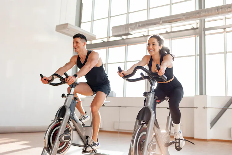 A man and a woman ride exercise bicycles in a gym.