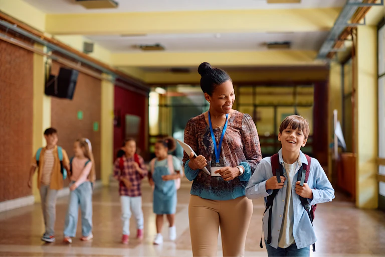 A woman walks down a hall smiling at a young boy next to her, with a group of children behind them in the hallway.