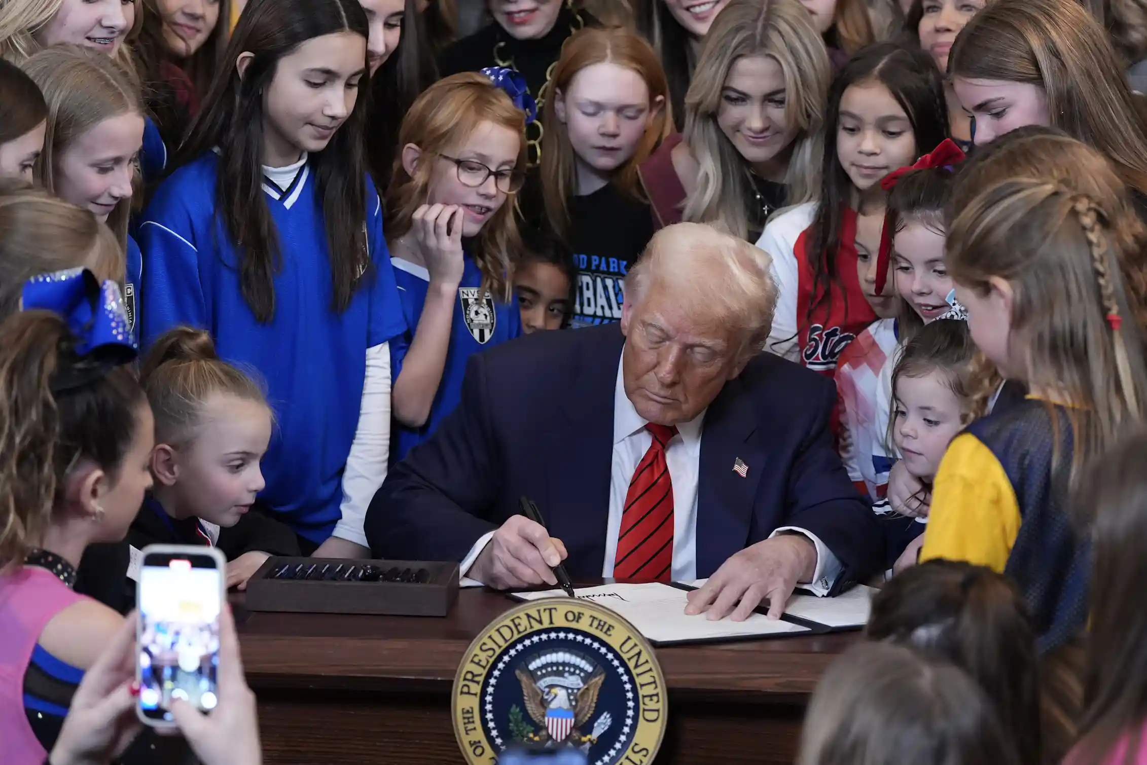 Donald Trump signs a document at a desk bearing the presidential seal, surrounded by girls and young women, some in sports jerseys