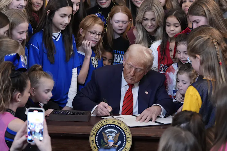 Donald Trump signs a document at a desk bearing the presidential seal, surrounded by girls and young women, some in sports jerseys