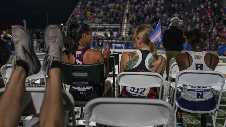 Seen from behind, track athletes sit in chairs in a stadium at night, one with a trans pride flag tucked in her hair.