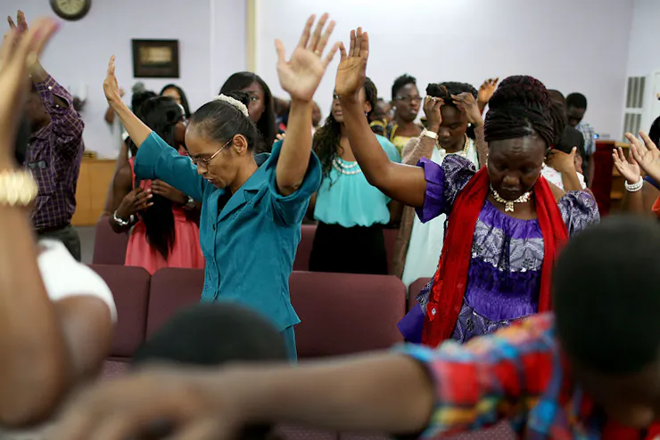 A small crowd of people stand and raise their arms in prayer as they look downward.
