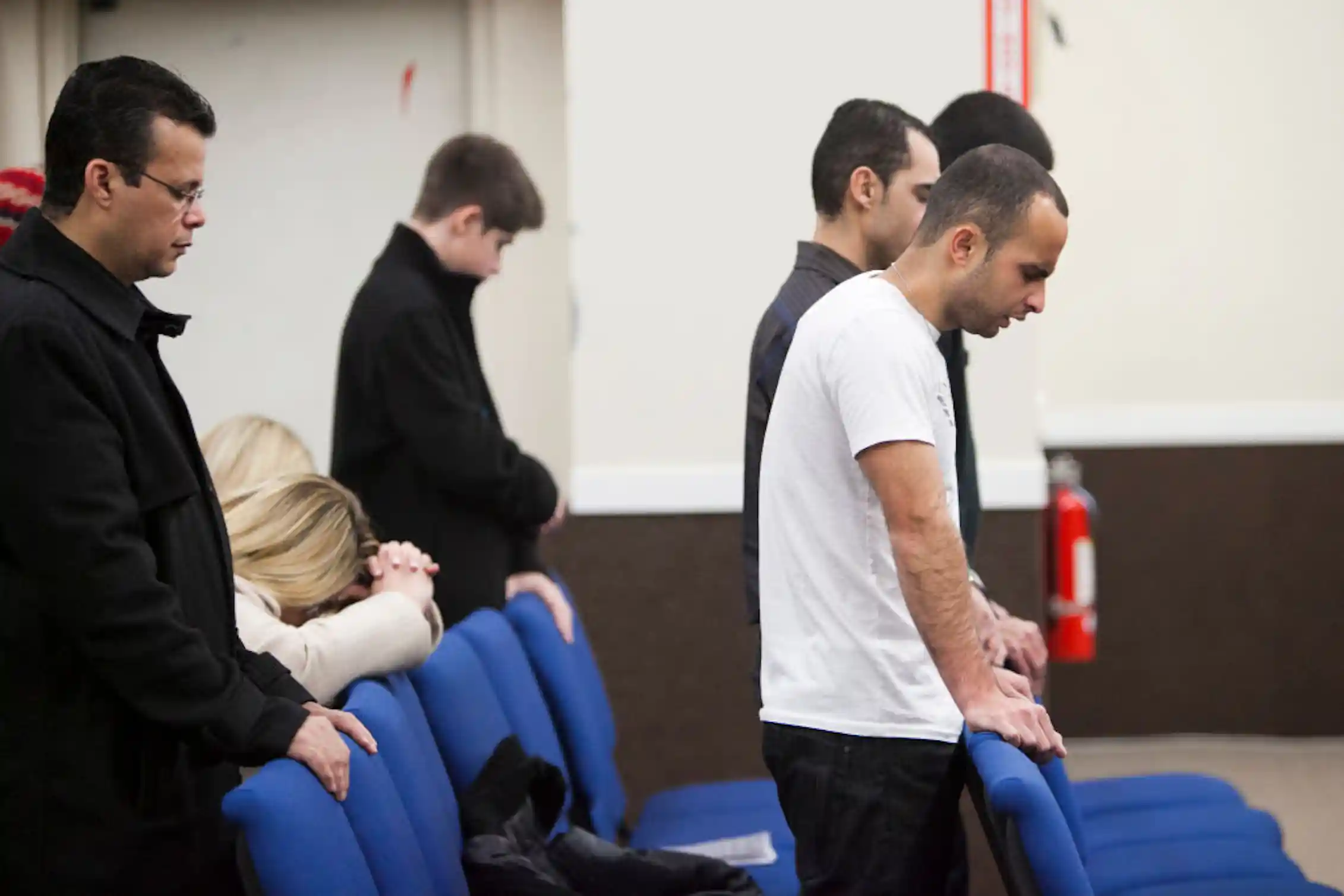 Several men bow their heads in prayer as they stand behind blue chairs.