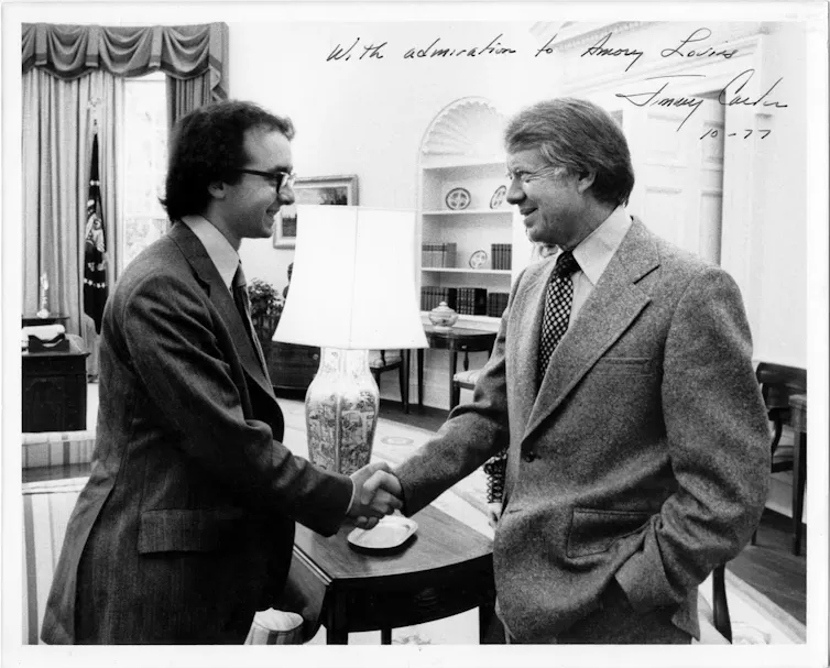 Two men in suits shake hands and smile.