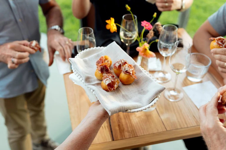 A platter of round rolls with cured meat stuffed into them, with a table, wine glasses and people in the background.