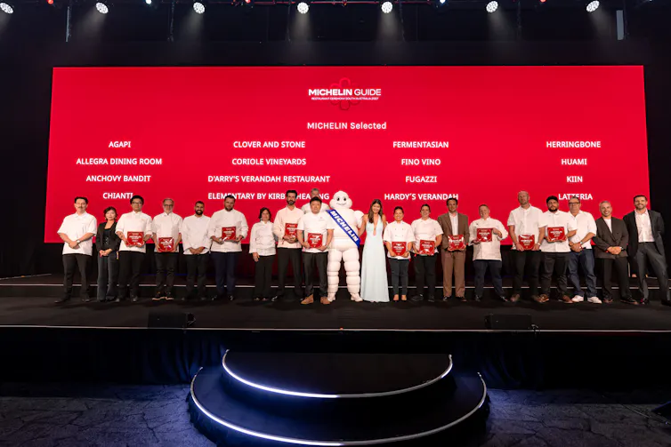 A large troup of people stands on stage with a Michelin mascot, against a screen with white text on red background.