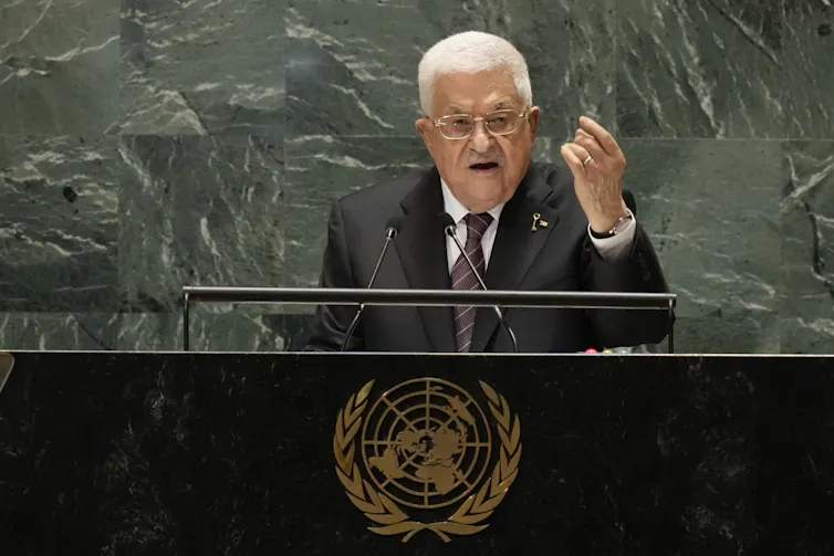 A white-haired man in a suit speaks from a podium.
