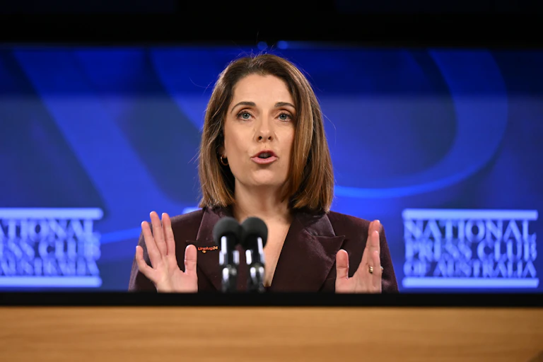 A woman with brown hairs speaks at a podium with a bright blue background.