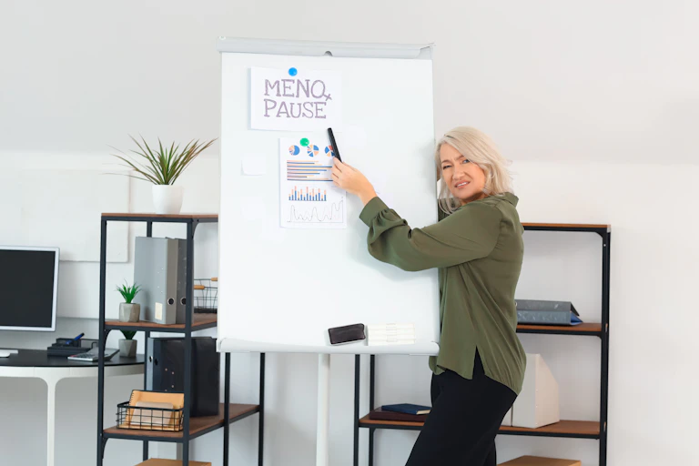 Une femme d'affaire est debout, à côté d'un tableau blanc, sur lequel est écrit "MENOPAUSE", mot qu'elle point avec un stylo. Des courbes et graphiques sont positionnés plus bas, dans le tableau.