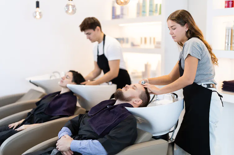 A young woman and young man both washing customers' hair at basins.