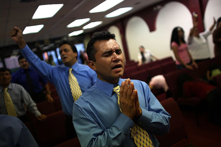 Two men wearing ties and blue button-up shirts, with their eyes closed in prayer, stand inside a room with rows of red seats.